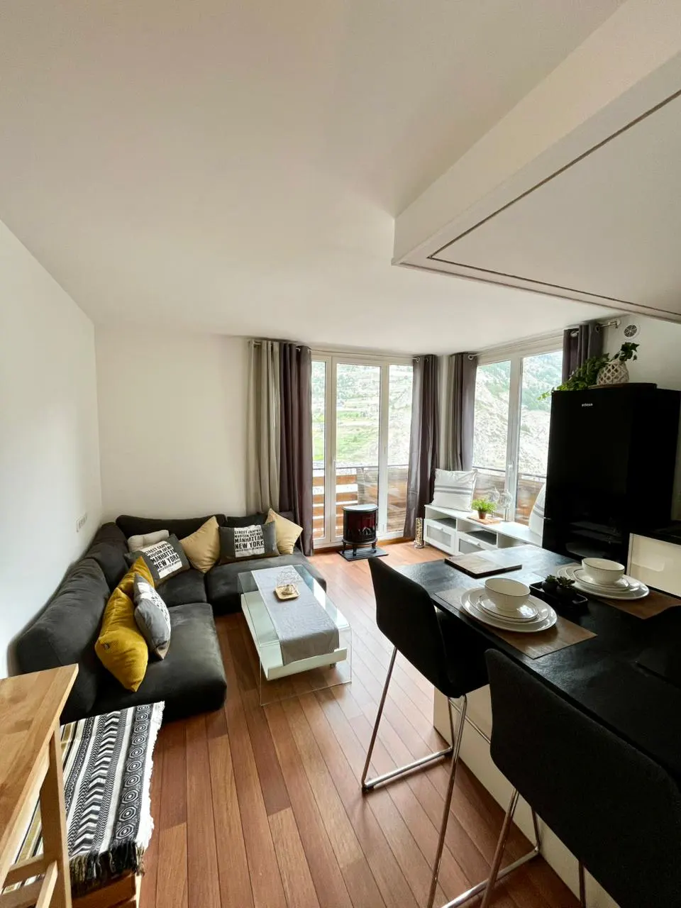 Open-concept living room with hardwood floors, a gray sectional sofa with pillows, and a glass coffee table. Balcony with mountain views.