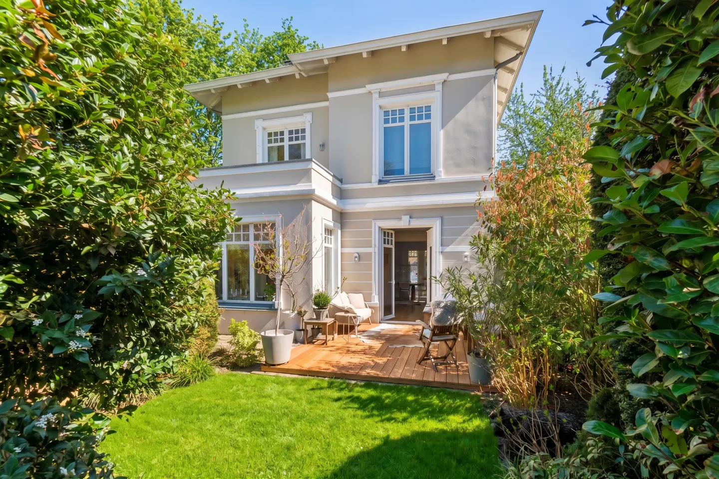 Two-story gray house with white trim, a wooden deck, and a green lawn surrounded by lush greenery.
