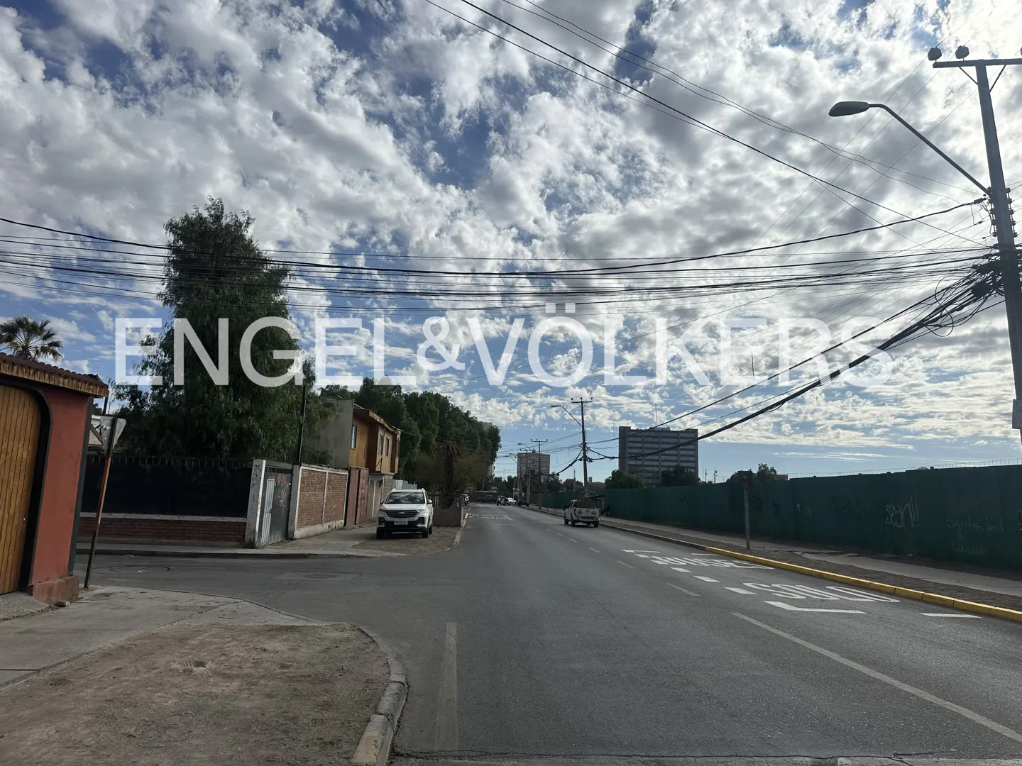 Street view with houses, cars, and a cloudy sky. Power lines crisscross above the road. Engel & Völkers logo is superimposed.