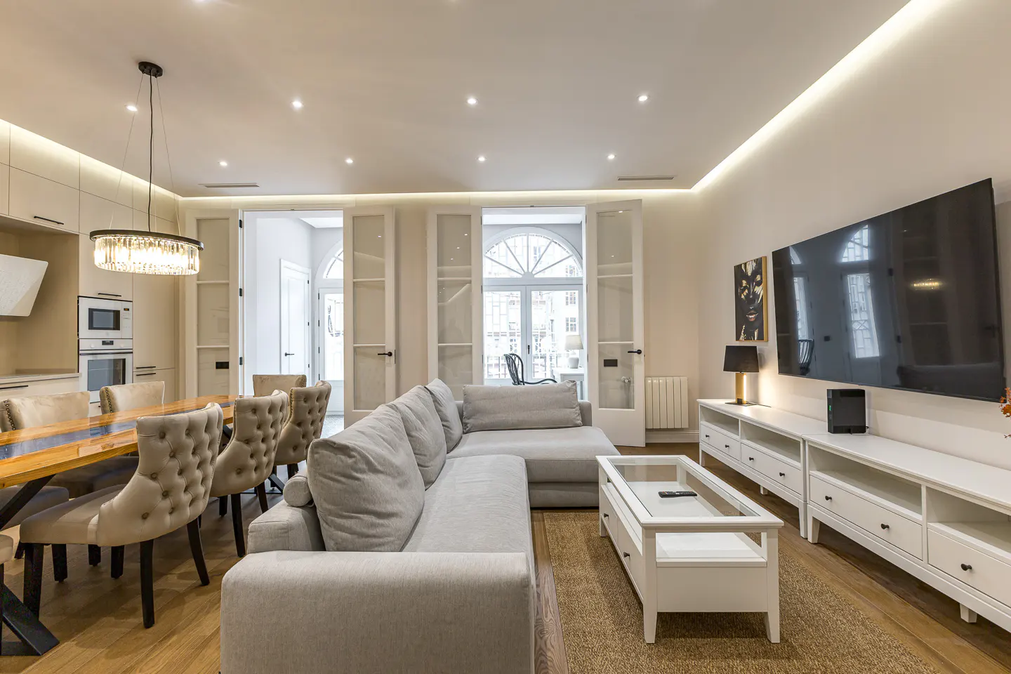 Bright, modern living room with a gray sectional sofa, white media console with a large TV, and a dining table with tufted chairs.