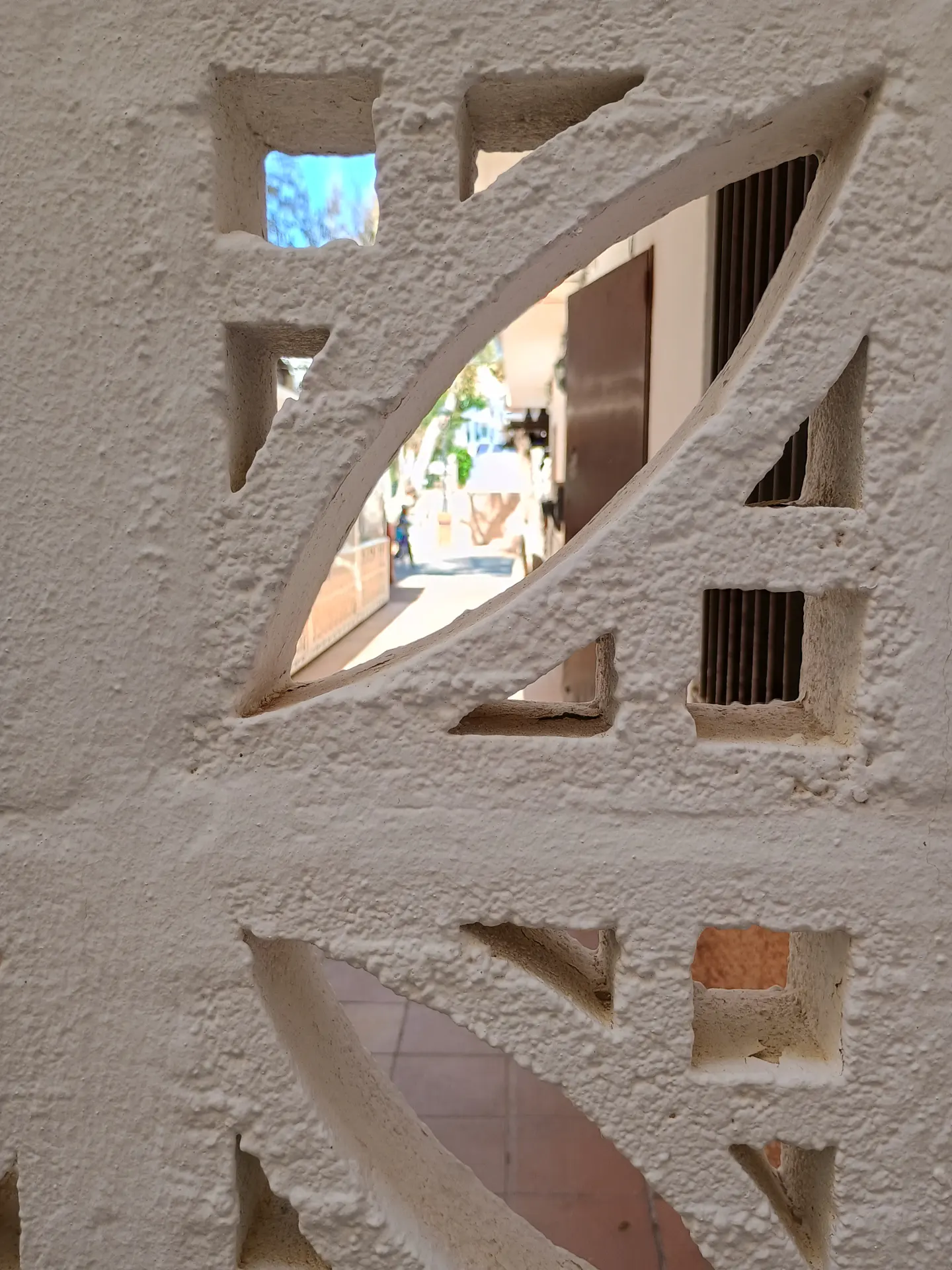 Close-up of a textured white wall with decorative cutouts revealing a glimpse of a courtyard and building beyond.