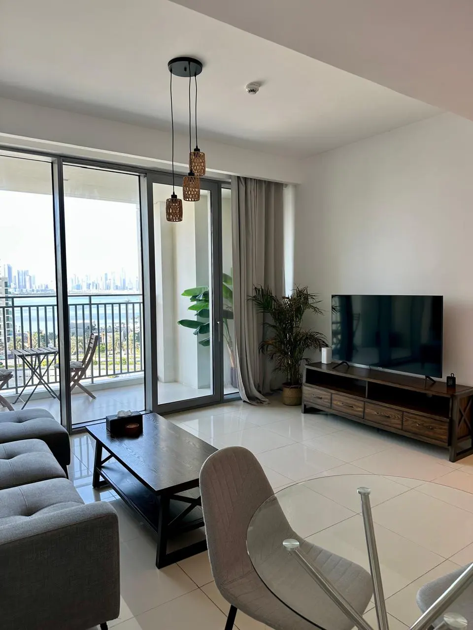 Bright living room with a gray sofa, dark wood coffee table, and a TV on a wooden stand. Balcony with ocean view.