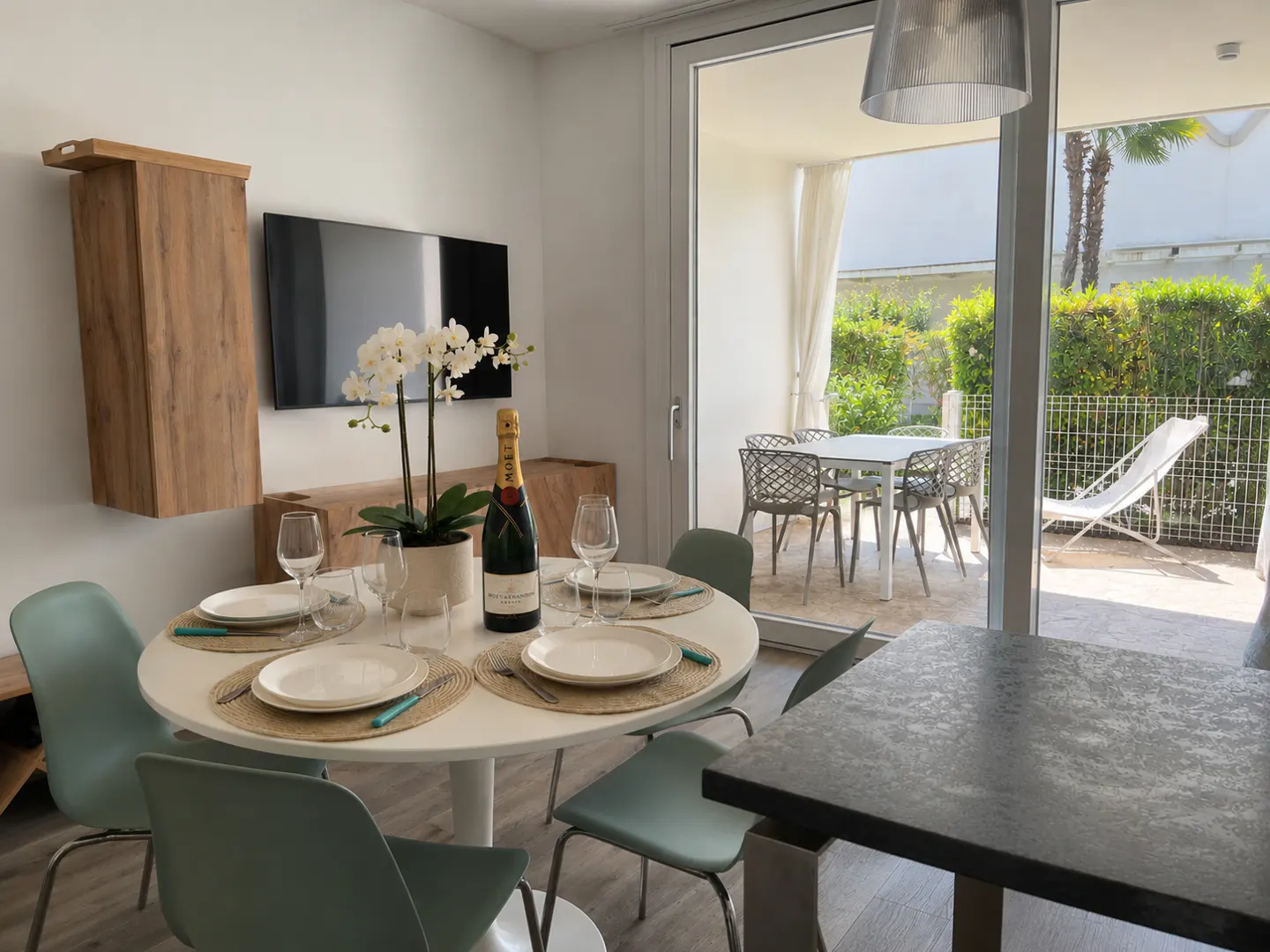 Bright dining area with a white table set for four, champagne, and orchid centerpiece. Patio with table and lounge chair visible through sliding glass doors.