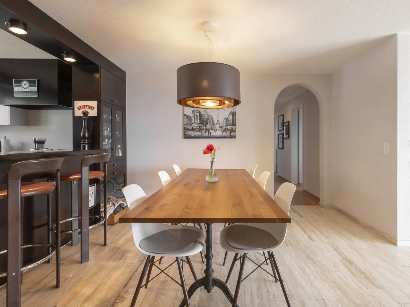 Dining area with a wooden table, white chairs, a modern pendant light, and a bar counter with stools in the background.