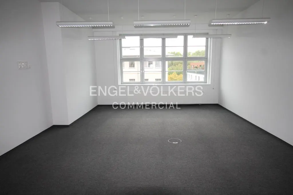 Empty office space with gray carpet, white walls, and a large window. Overhead lighting fixtures illuminate the room. Engel & Volkers logo is visible.