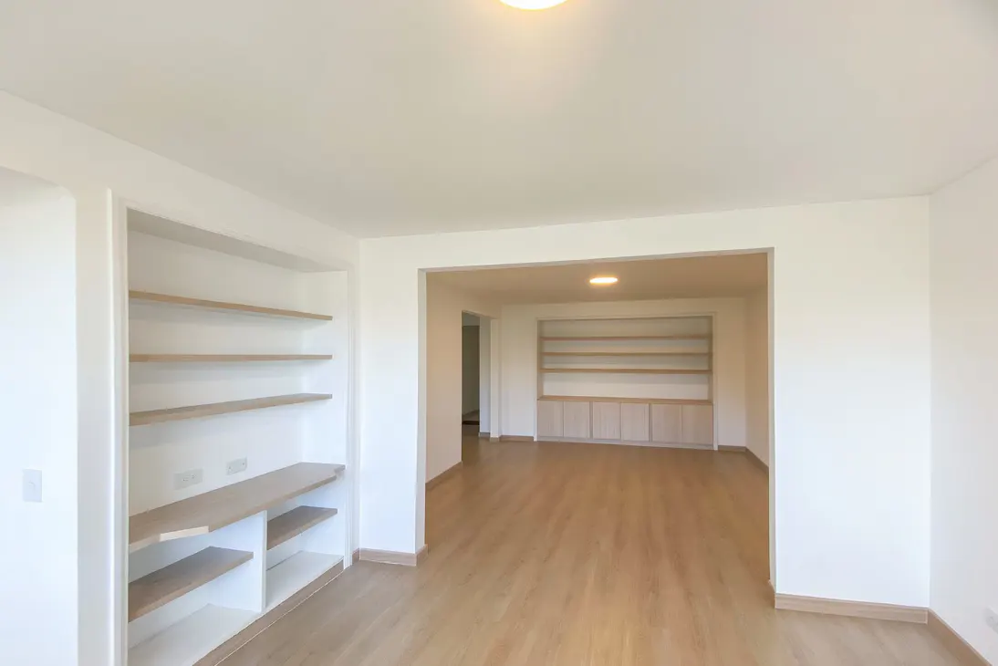 Bright, empty room with light wood floors and white walls. Built-in shelves are on the left and in the adjacent room.
