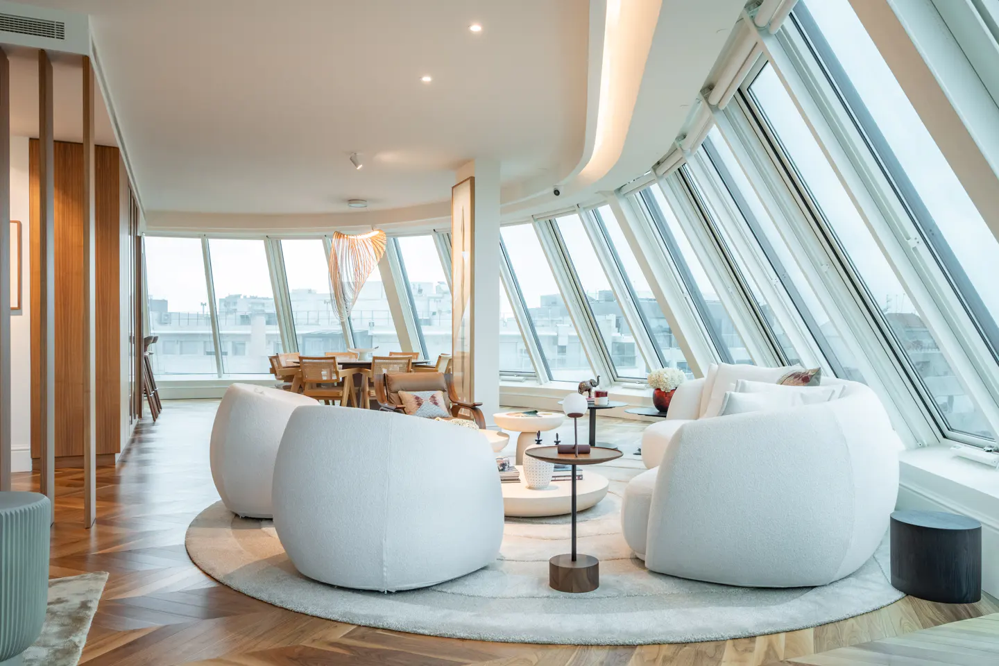 Bright, modern living room with white chairs, wood floors, and large windows overlooking a city.