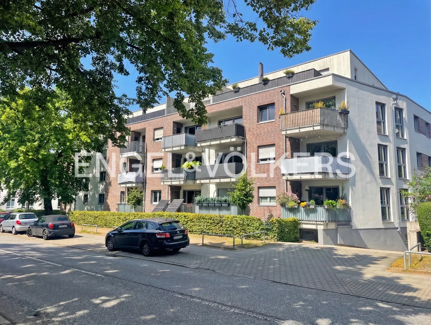 Modern apartment building with brick and white facade, balconies, and parked cars on a sunny street with green trees.