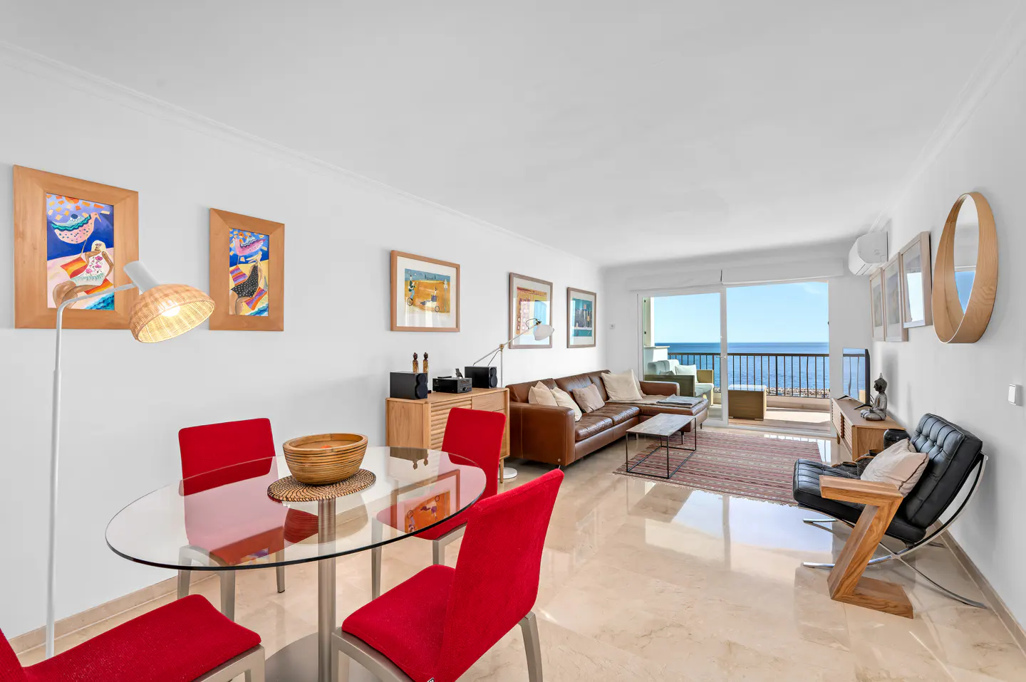 Bright living room with ocean view. Red chairs surround a glass table. A brown leather sofa and black chair sit on a marble floor.
