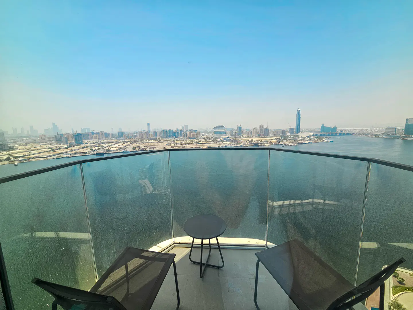 Balcony view with two chairs and a table overlooking a city skyline and blue water on a sunny day.