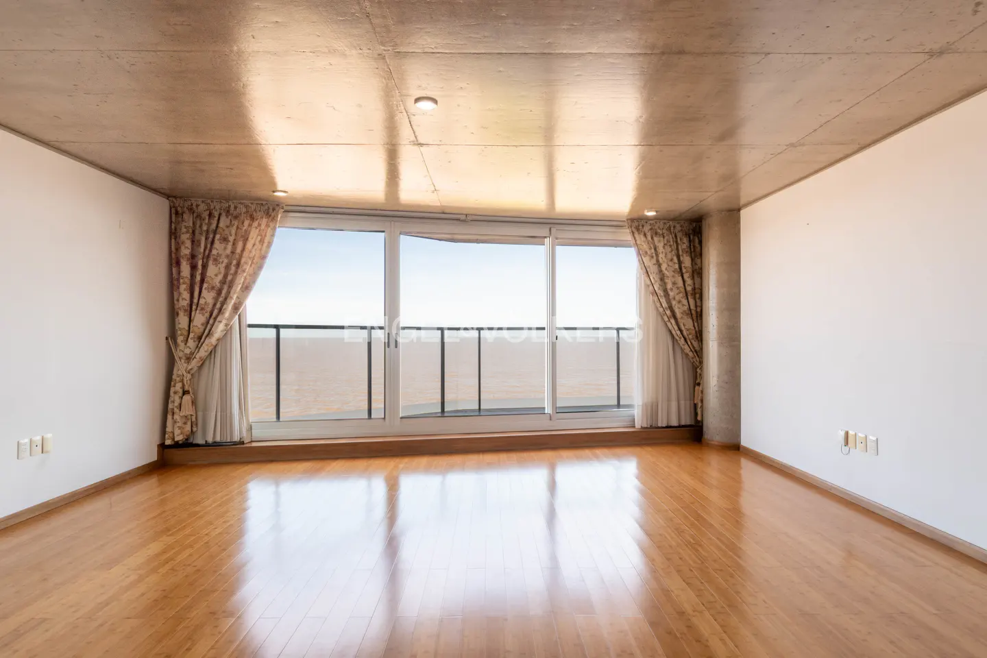 Empty room with hardwood floors, white walls, and a concrete ceiling. Large windows with curtains overlook a balcony and the ocean.