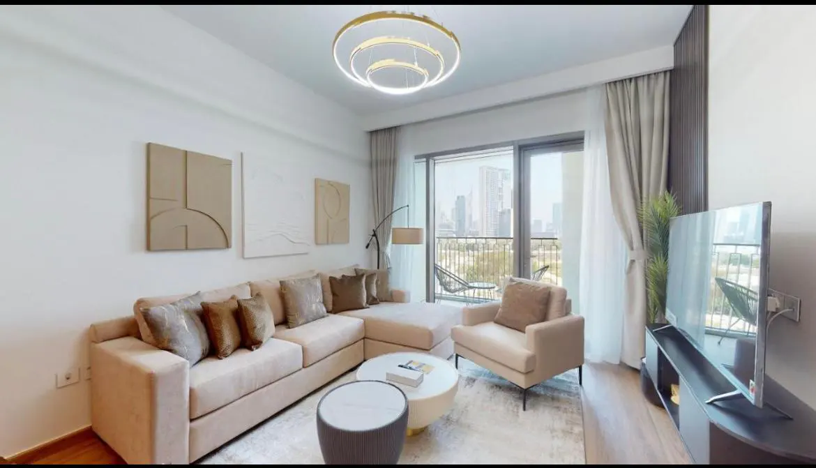 Living room with beige sectional sofa, chair, and city view. Modern light fixture and neutral artwork add to the bright, airy space.