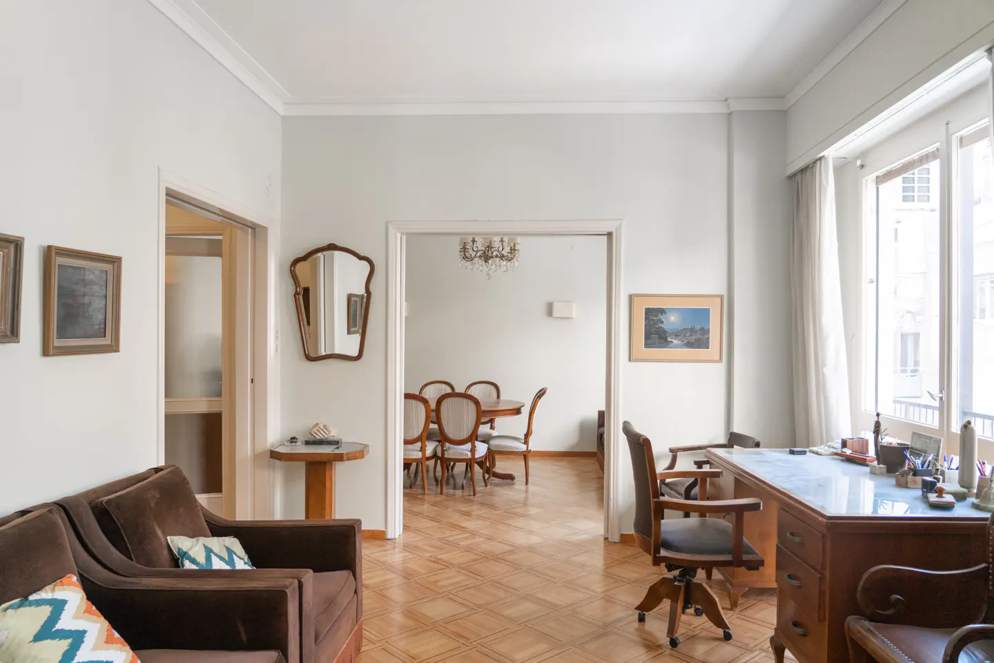 Interior view of a living room with brown sofa, desk, and dining room visible through an open doorway. Parquet flooring and light gray walls.