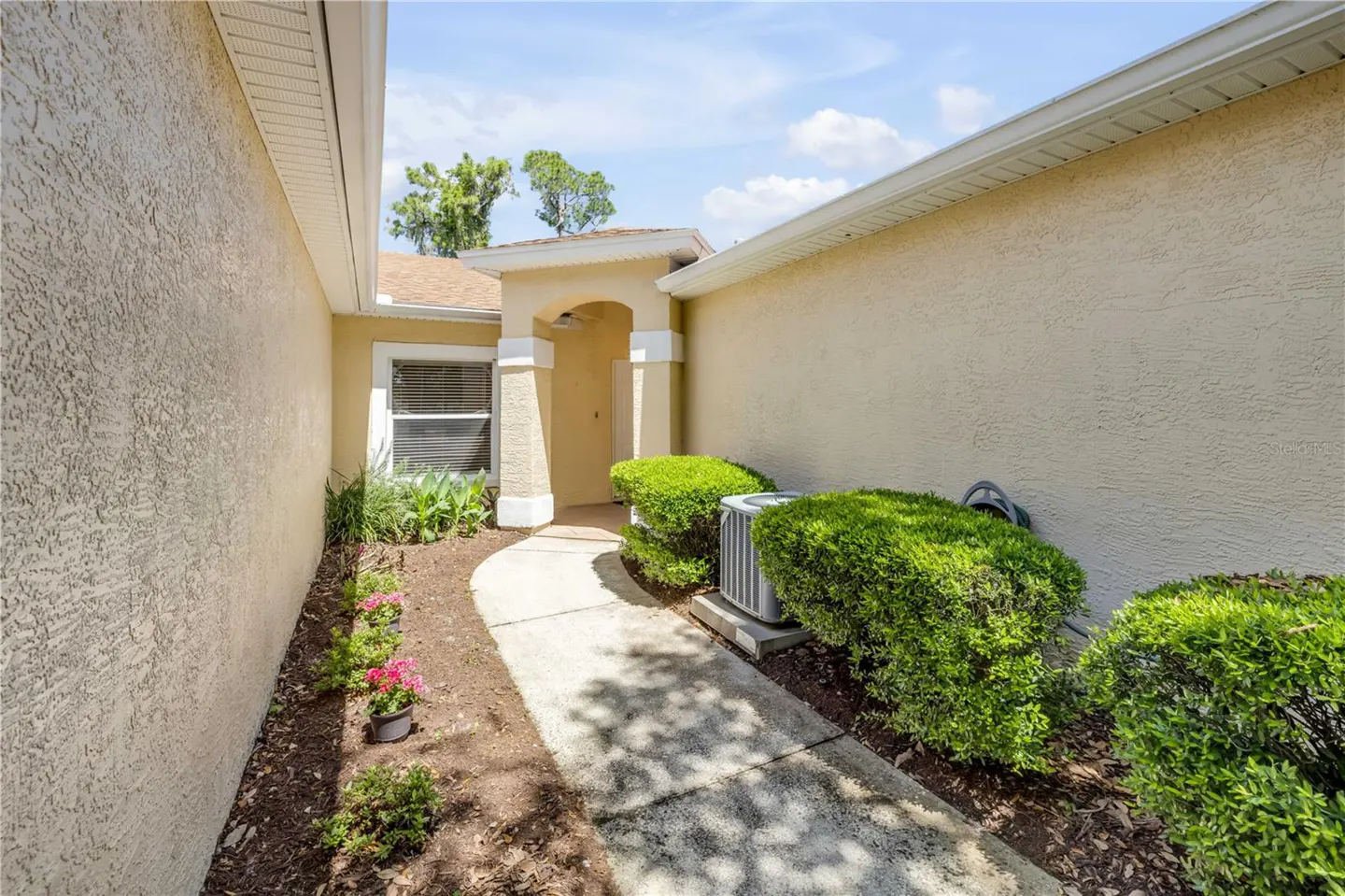 Exterior view of a beige house with a curved walkway leading to the front door, flanked by green bushes and flowerbeds.
