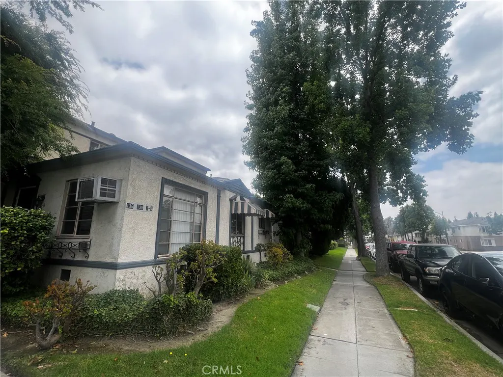 Exterior view of a light beige stucco apartment building with dark trim, a sidewalk, and parked cars on the street.