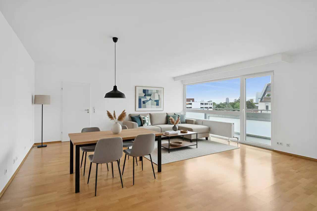 Bright, modern apartment interior with wood floors, dining table, and sectional sofa near a large window with a city view.