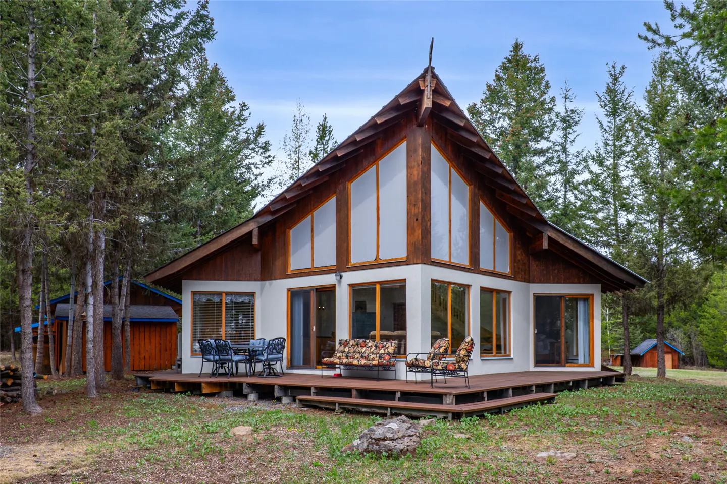 A-frame house with a wood deck, surrounded by tall trees. The house has large windows and a white exterior. Outdoor furniture is on the deck.