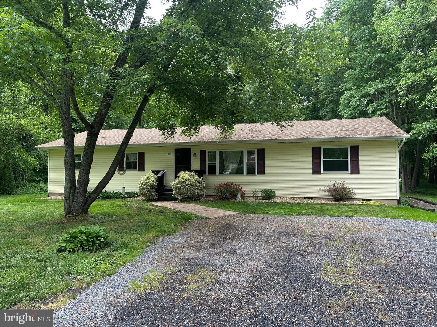 A one-story yellow house with brown shutters and a gravel driveway is surrounded by green trees.