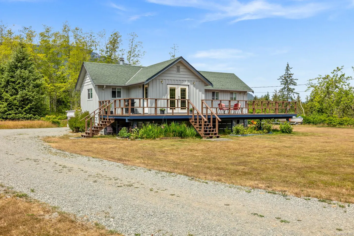 Exterior view of a one-story house with a green roof and a large wooden deck. A gravel driveway leads to the house.