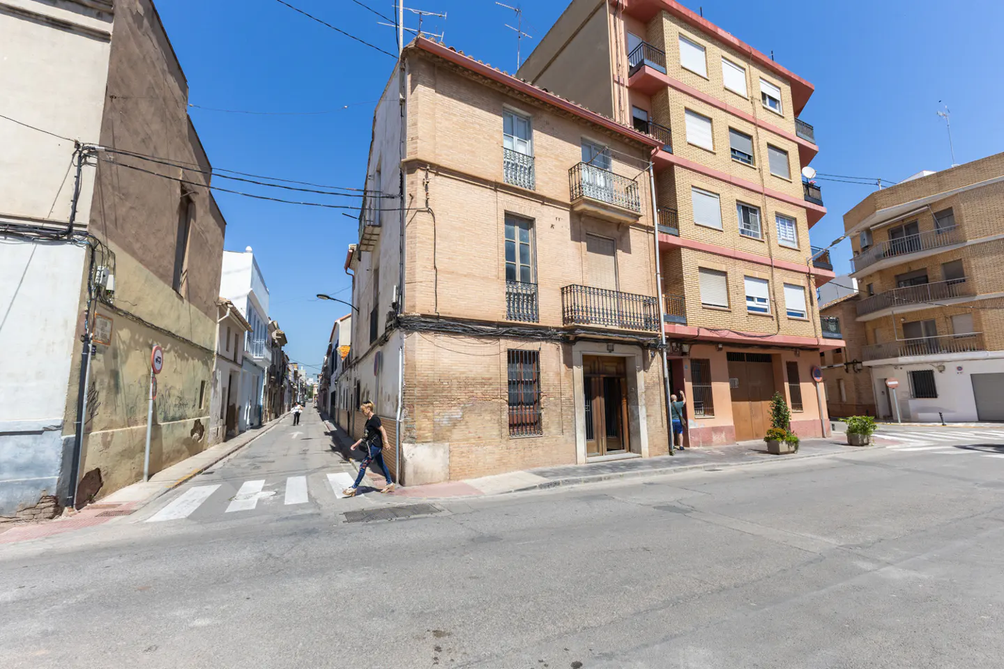 Street view of a tan brick building with balconies, a woman crossing the street, and other buildings under a clear blue sky.
