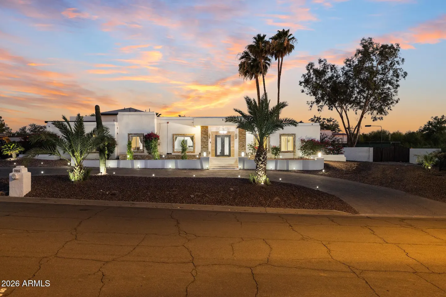 Single-story white home at sunset with palm trees, desert landscaping, and a curved driveway illuminated by path lights.