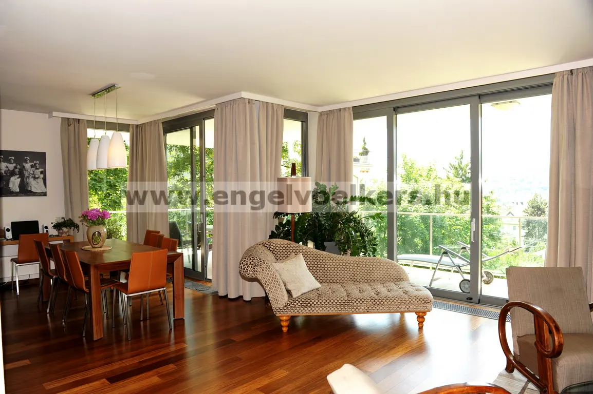 Bright living room with hardwood floors, dining table, and chaise lounge. Large windows offer a view of greenery.