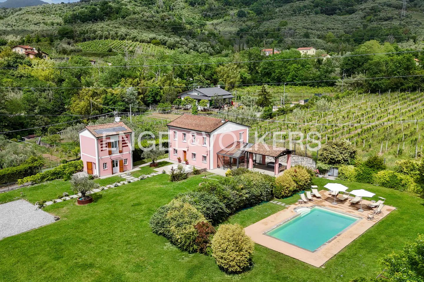 Aerial view of a pink house with a pool, lawn, and vineyard in the background.
