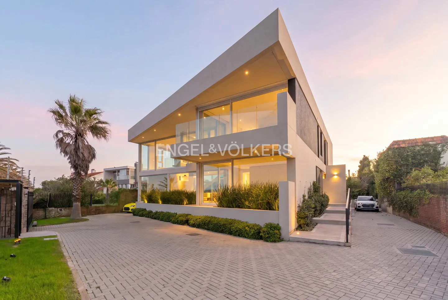 Modern two-story home with angular roof, floor-to-ceiling glass walls, and paved driveway at sunset.