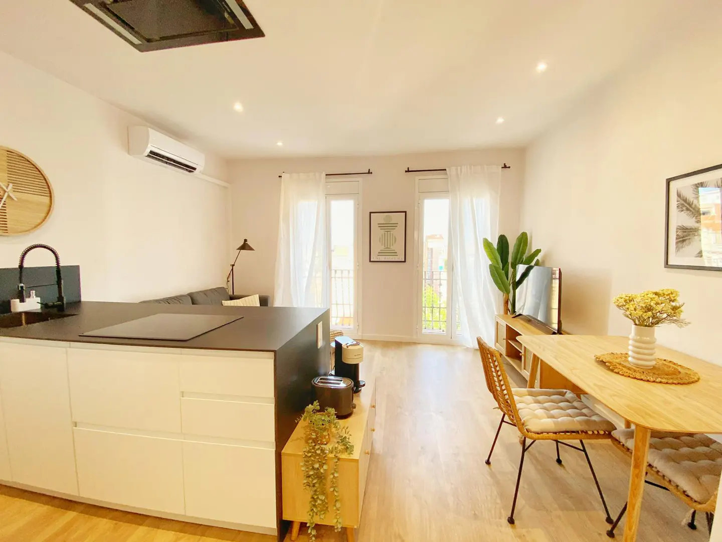 Bright, modern apartment interior with a kitchen island, dining table, and living area. White walls and light wood floors create a spacious feel.