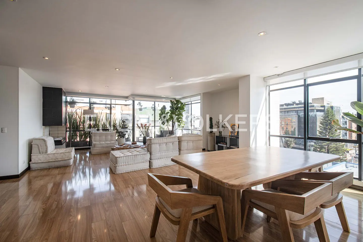 Bright, open-concept living space with wood floors, a dining table, and modular seating near large windows with city views.