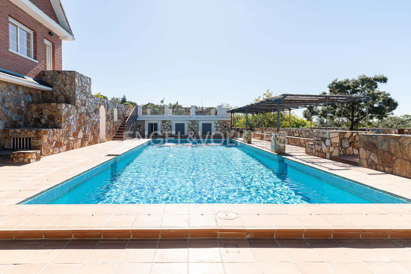 A rectangular blue tiled pool is surrounded by stone walls and a pergola, under a clear blue sky.