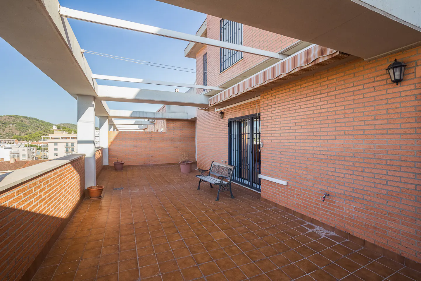 Wide shot of a brick patio with a bench, potted plants, and a pergola. A brick building is in the background.
