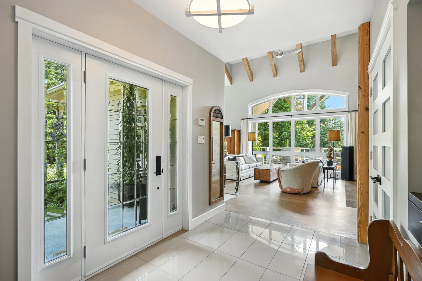 Bright foyer with white tile floor, glass paneled front door, and view into a living room with large windows.