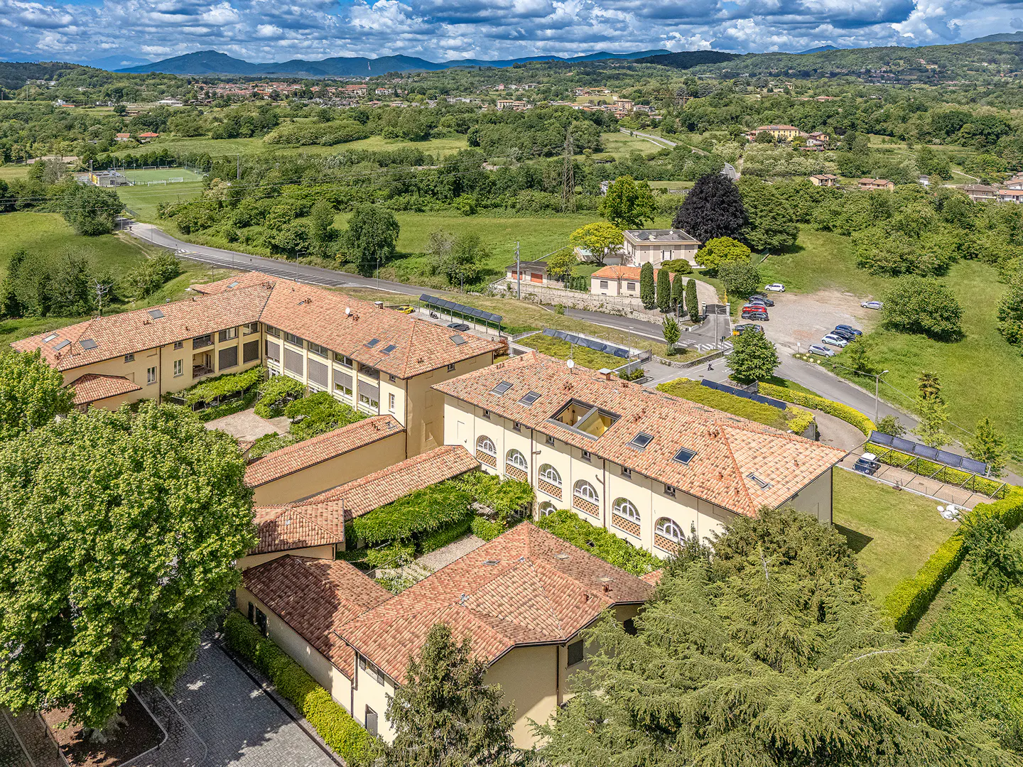 Aerial view of a large, tan building with a red tile roof, surrounded by green trees and grass. Mountains are visible in the background.