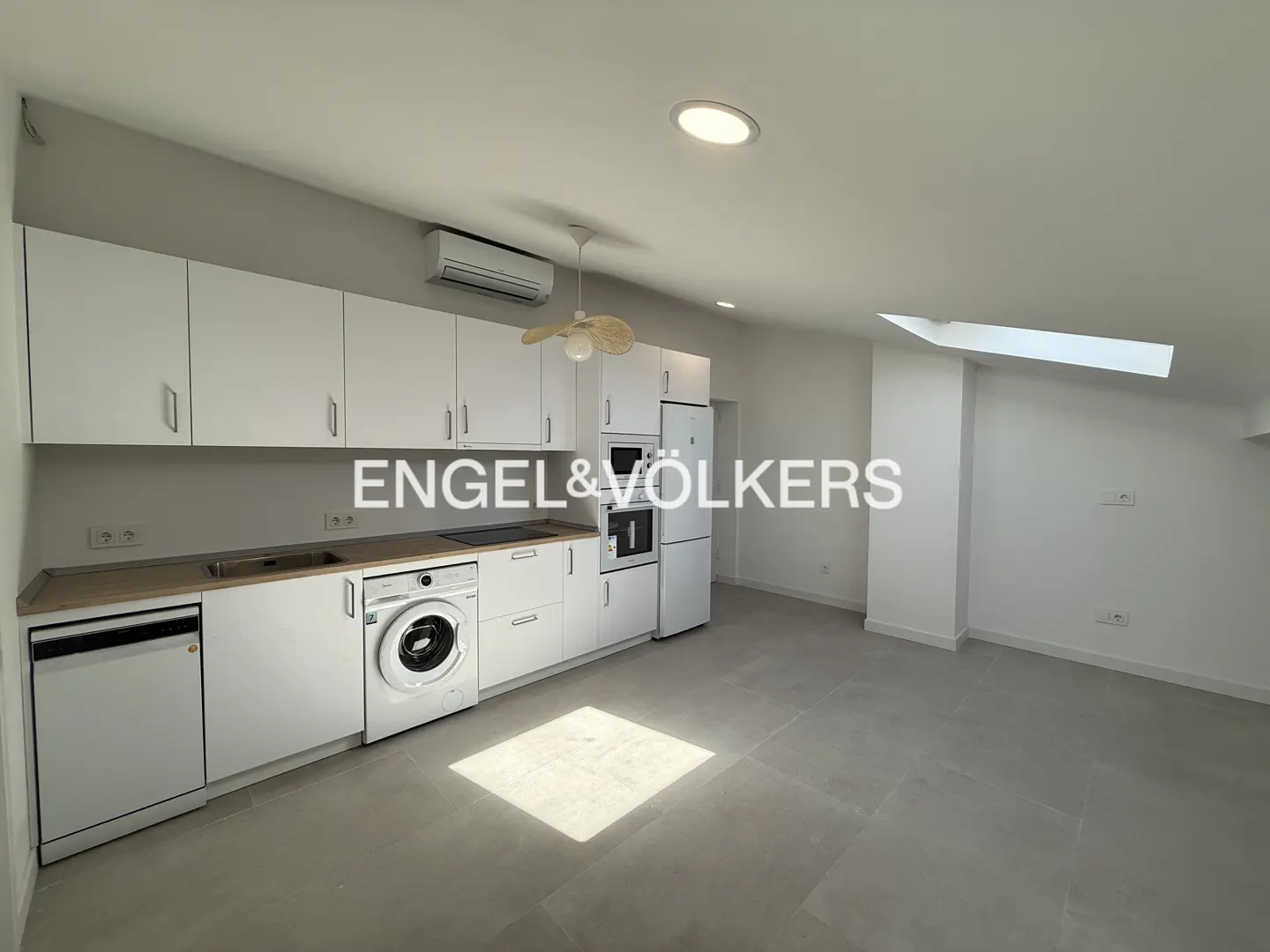 Bright, modern kitchen with white cabinets, appliances, and gray tile floor. A skylight provides natural light. Engel & Völkers logo overlay.