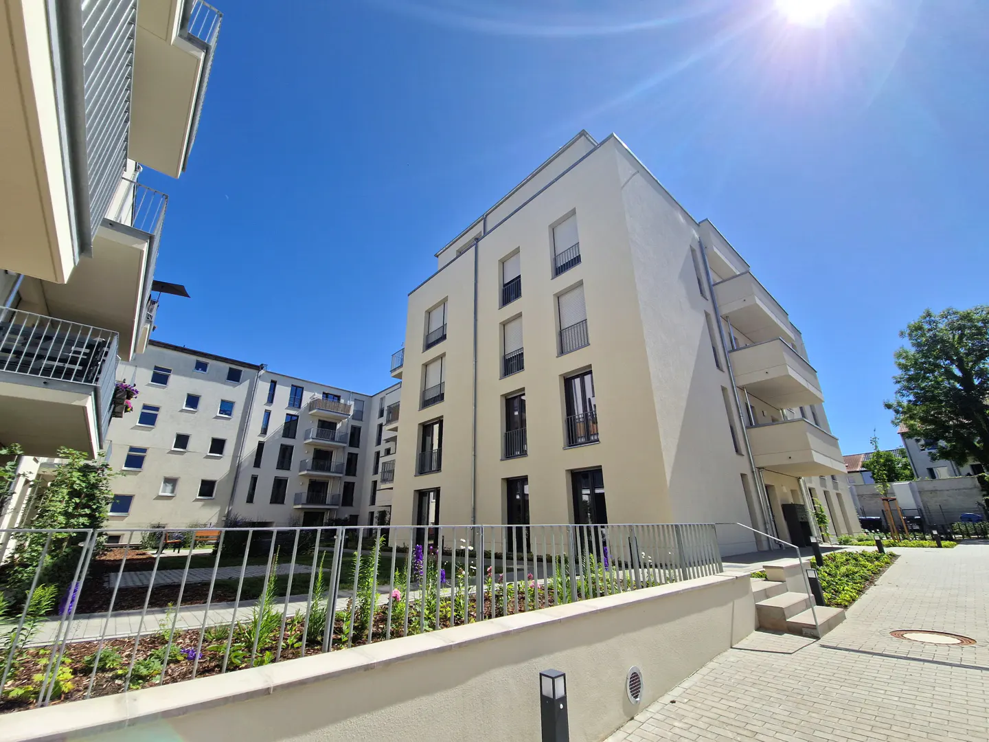 Exterior view of a modern, multi-story apartment building with balconies under a bright blue sky. A landscaped garden and paved walkway are in the foreground.