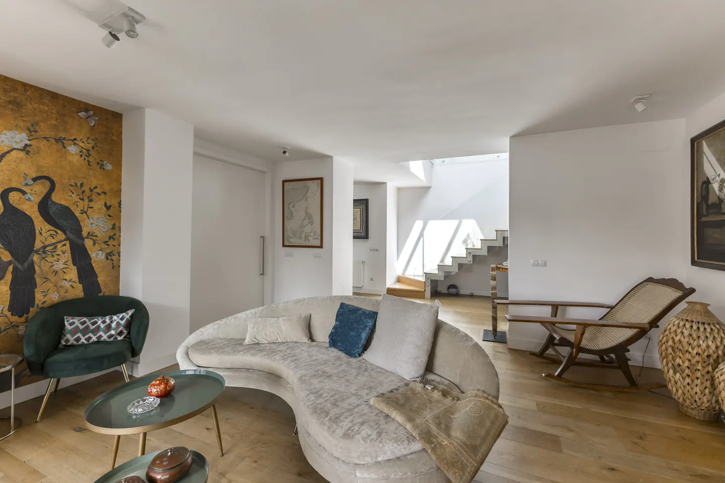 Bright living room with hardwood floors, a curved sofa with pillows, and a gold wall mural with birds. A rocking chair sits near stairs.