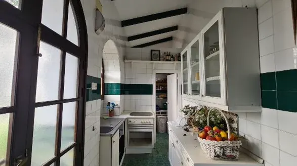 A narrow kitchen with white tile walls and dark green accents. White cabinets line one wall, and a basket of fruit sits on the counter. A dark wood-framed window is on the left.