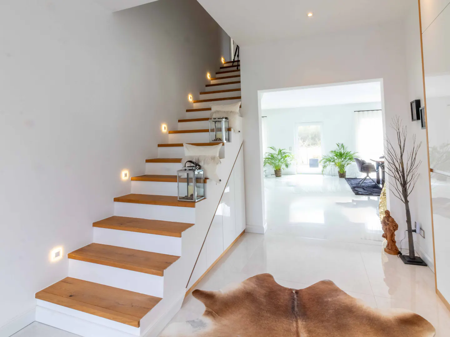 Bright, modern entryway with wood and white stairs, lit by wall sconces. A cowhide rug sits on the white tile floor. View into a sunlit living room.