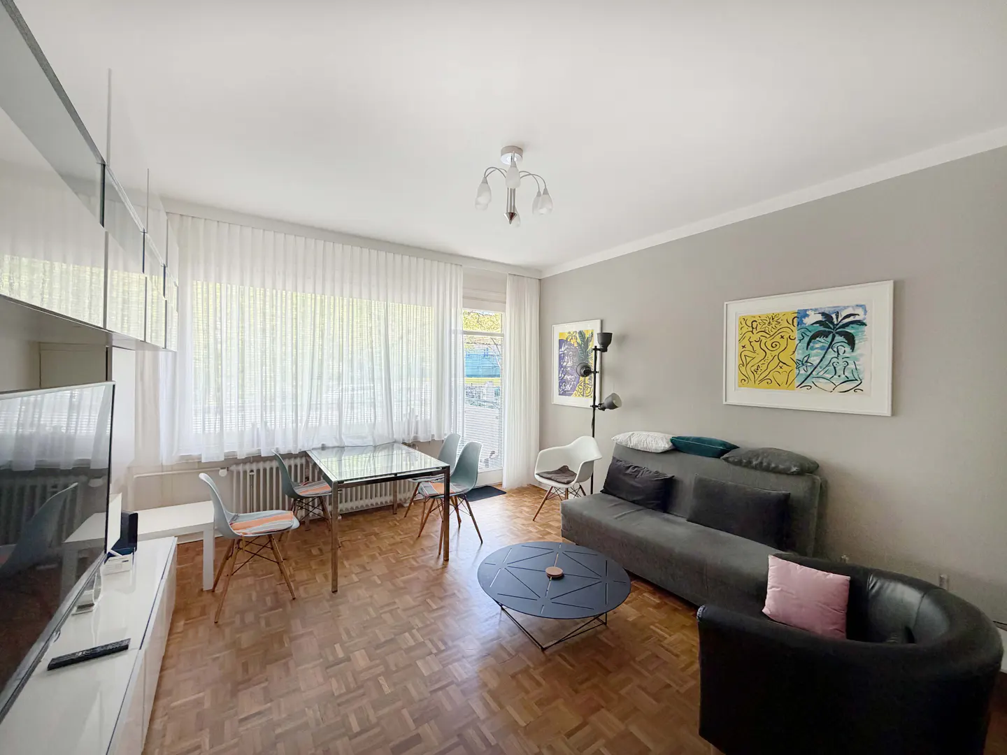 Bright living room with parquet floors, gray walls, and white trim. A glass table with chairs sits near a window with sheer curtains. A gray sofa and black chair are near colorful artwork.