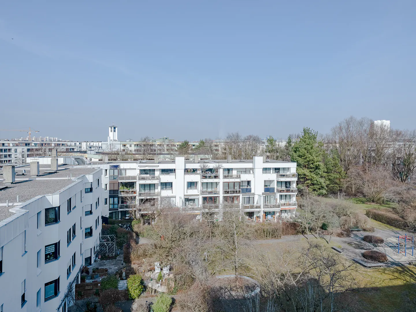 View of white apartment buildings with balconies, surrounded by trees and a playground under a clear blue sky.