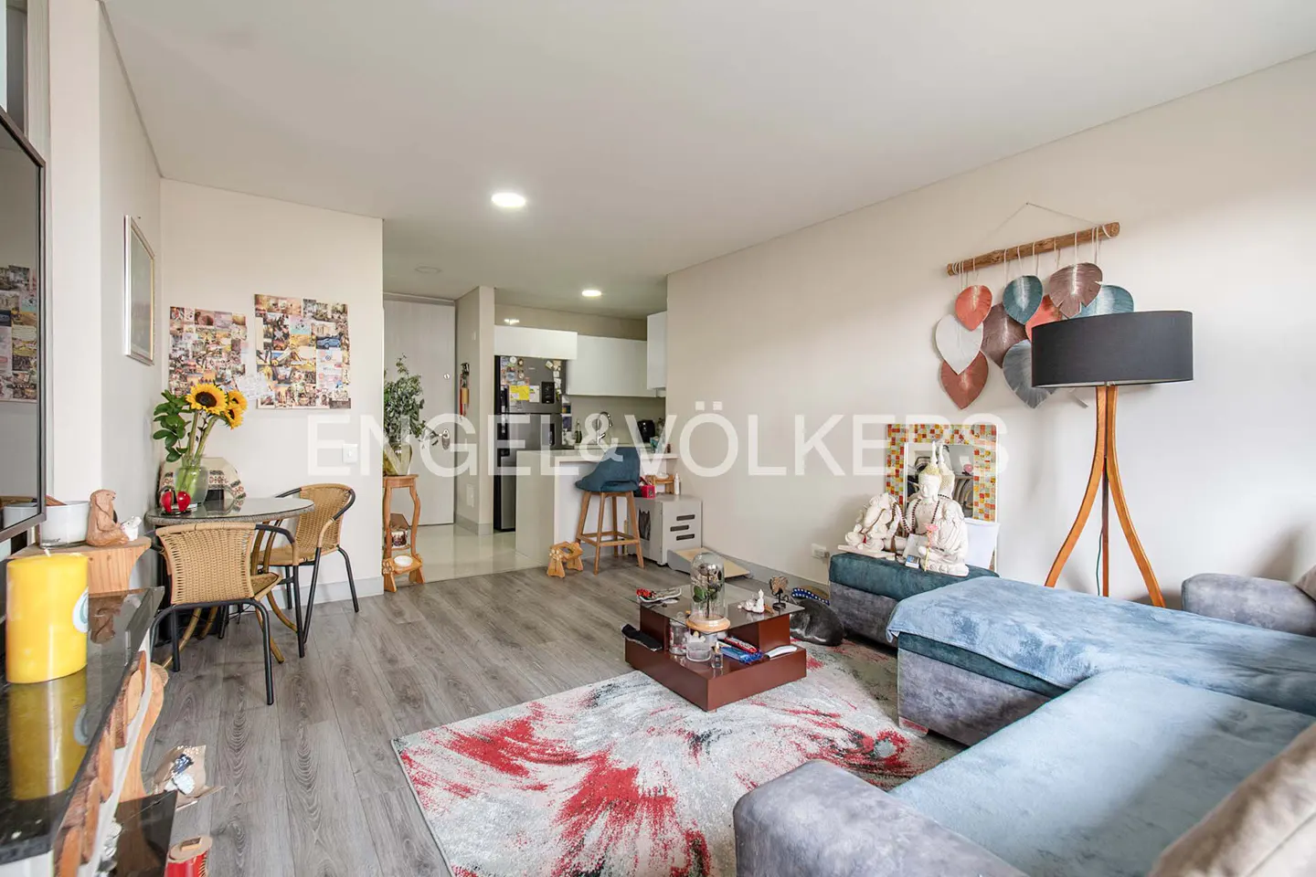Bright living room with gray wood floors, a blue sectional sofa, and a red and gray patterned rug. A dining table with sunflowers sits near a wall of photos.