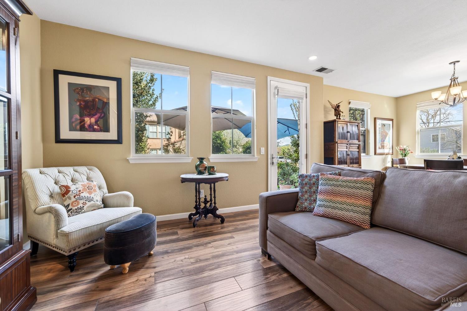 Living room with hardwood floors, tan walls, a gray sofa, and a tufted chair with a floral pillow. Windows overlook a patio.