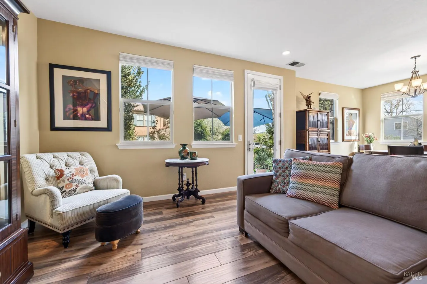 Living room with hardwood floors, tan walls, a gray sofa, and a tufted chair with a floral pillow. Windows overlook a patio.