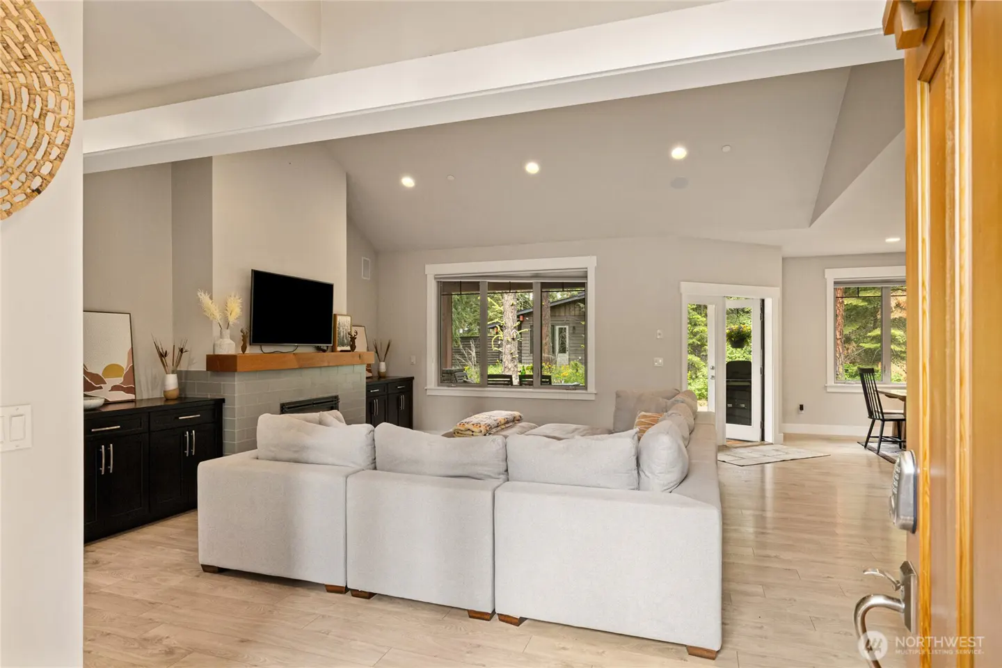 Open-concept living room with a large white sectional sofa, fireplace, TV, and light wood floors. Windows offer views of the outdoors.