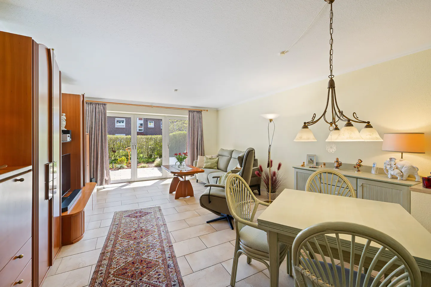 Bright living room with dining area, light tile floors, and a view of the garden through glass doors. A brown rug adds warmth.