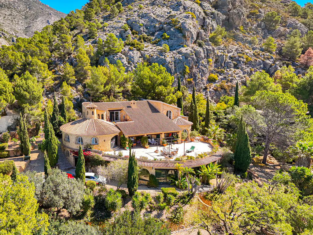 Aerial view of a tan stone house with a brown tile roof, surrounded by green trees and a rocky mountain.