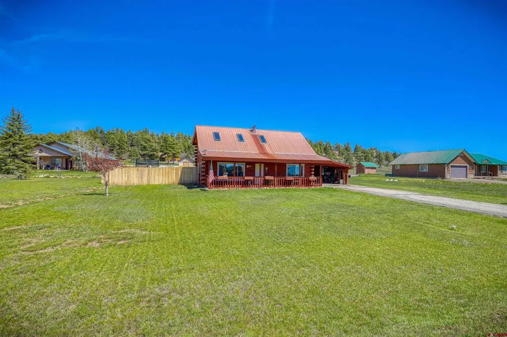 Exterior view of a red log cabin-style house with a metal roof, surrounded by a green lawn and trees under a clear blue sky.
