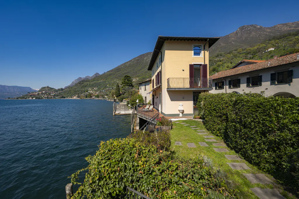 Lakefront property with a yellow house, balcony, and stone path leading to a green hedge. Mountains and blue sky in the background.