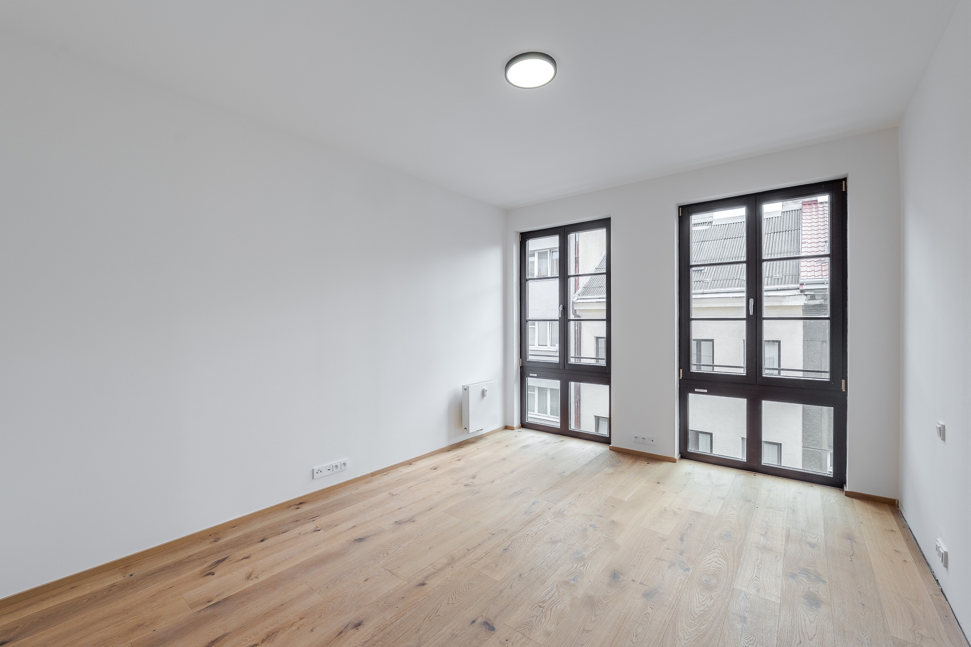 Empty room with white walls, light wood flooring, and two large windows with dark frames overlooking a city building. [AI-generated text]